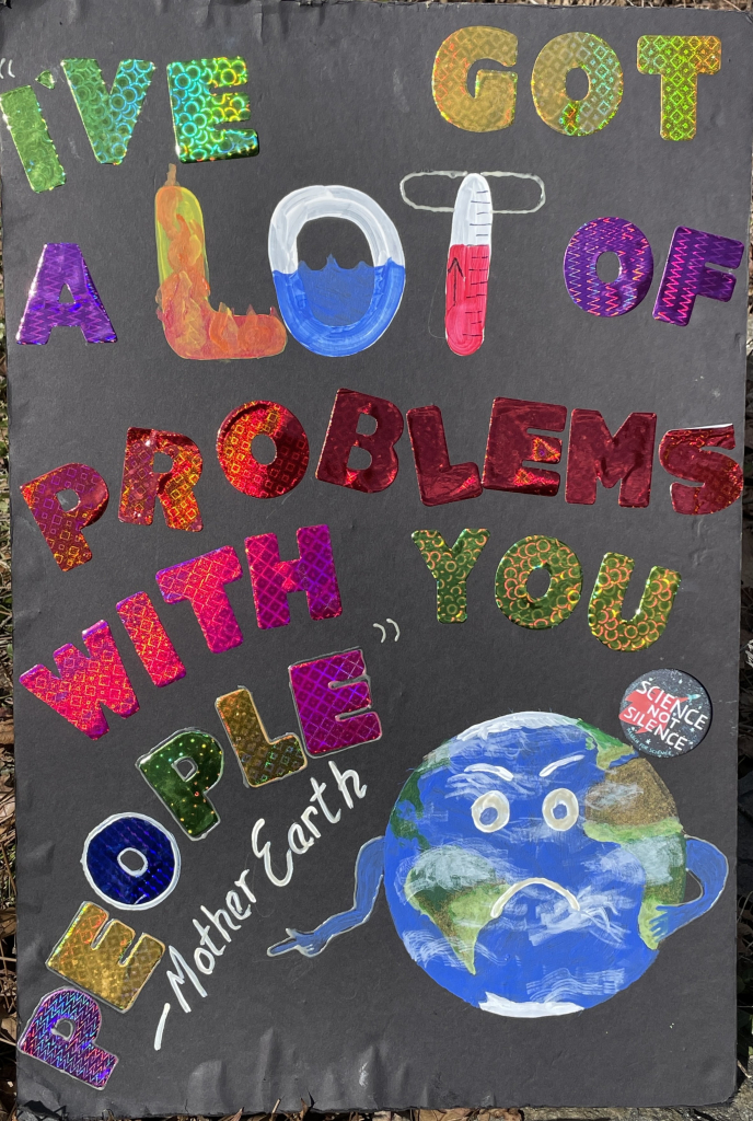 A colorful protest sign on a black background featuring the text "I've got a lot of problems with you people" attributed to "Mother Earth." The sign includes a cartoon depiction of the Earth with arms and a frown, alongside images of flames, water, and a rising thermometer. A button attached says “Science not Silence” from the 2017 March for Science in a protest against cuts to science funding made during the last time we were here. 