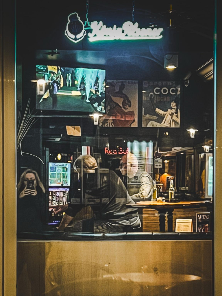 Gäste in einer Bar durch das Fenster fotografiert. Die Szene ist eher trist. Oben links läuft ein Fernseher, aber keiner schaut hin. Ein Gast sitzt an der Bar, ein anderer schräg davor vor einem Spielautomaten. Oben im Fenster Leuchtreklame mit König Pilsener. Es spiegeln sich diverse gegenüberliegende Fenster. 

Spezialeffekt: ich habe beim spontanen Fotografieren nicht darauf geachtet und habe mich als Spiegelung prominent mit fotografiert 

Guests in a bar photographed through the window. The scene is rather dreary. There's a TV at the top left, but no one is looking. One guest sits at the bar, another side by it in front of a slot machine. At the top of the window neon sign with King Pilsener. Various opposite windows are reflected.

Special effect: I didn't pay attention to it when photographing spontaneously and photographed myself prominently as a reflection