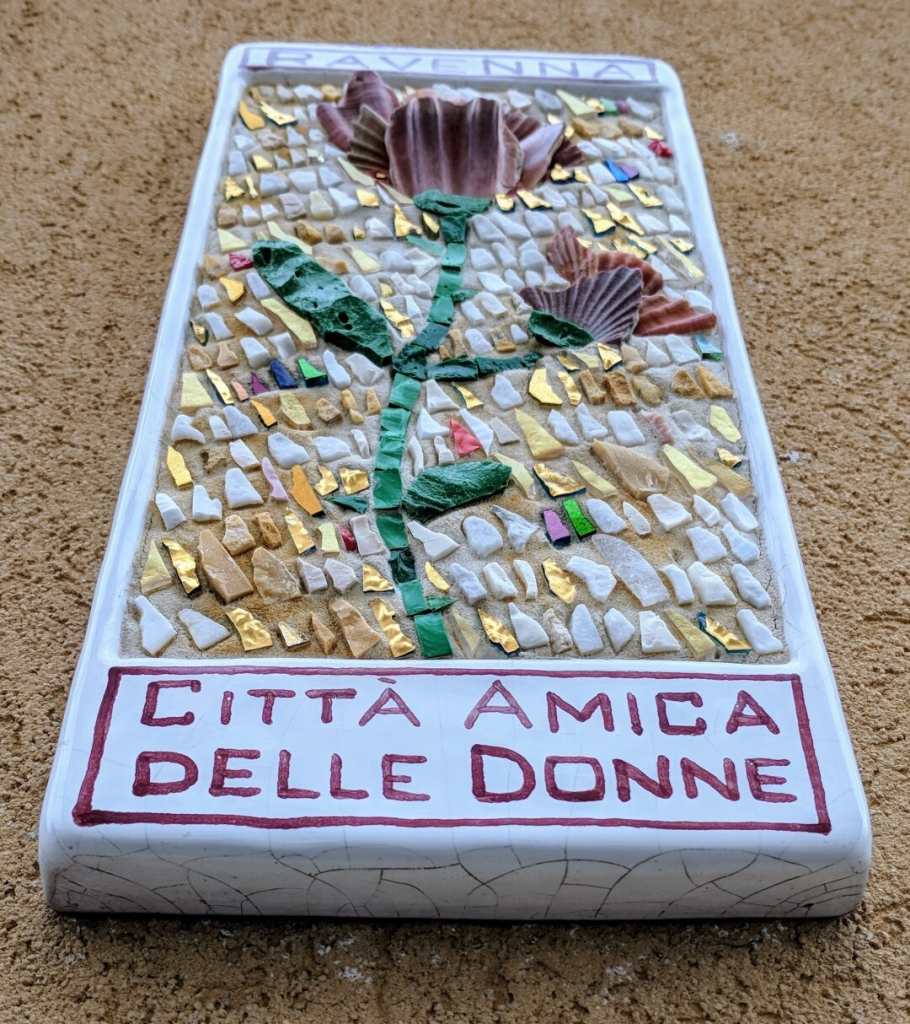 A slightly angled close-up shot of a rectangular, mosaic-decorated plaque lying on a textured, sandy surface. The plaque has a white background with a decorative border and features a stylized flower made from mosaic pieces in shades of brown, green, gold, and other colours. Below the flower is the text "CITTA AMICA DELLE DONNE" in capital letters, painted in a dark colour within a white rectangle outlined in red. At the top edge, partially visible, is the word "RAVENNA".