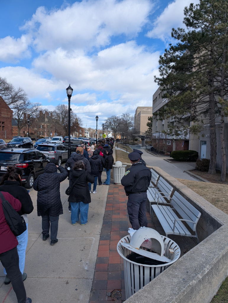 A very long line of people waiting to be admitted on the sidewalk and steps in front of Malden High School