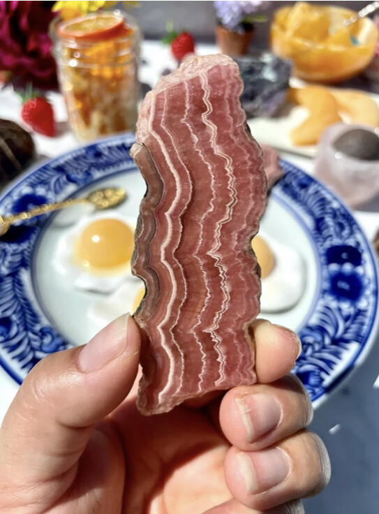 A hand holds a long thin piece of rhodochrosite which looks remarkably like a piece of bacon. In the background is a breakfast table, complete with two fried eggs on a plate and assorted fruit.