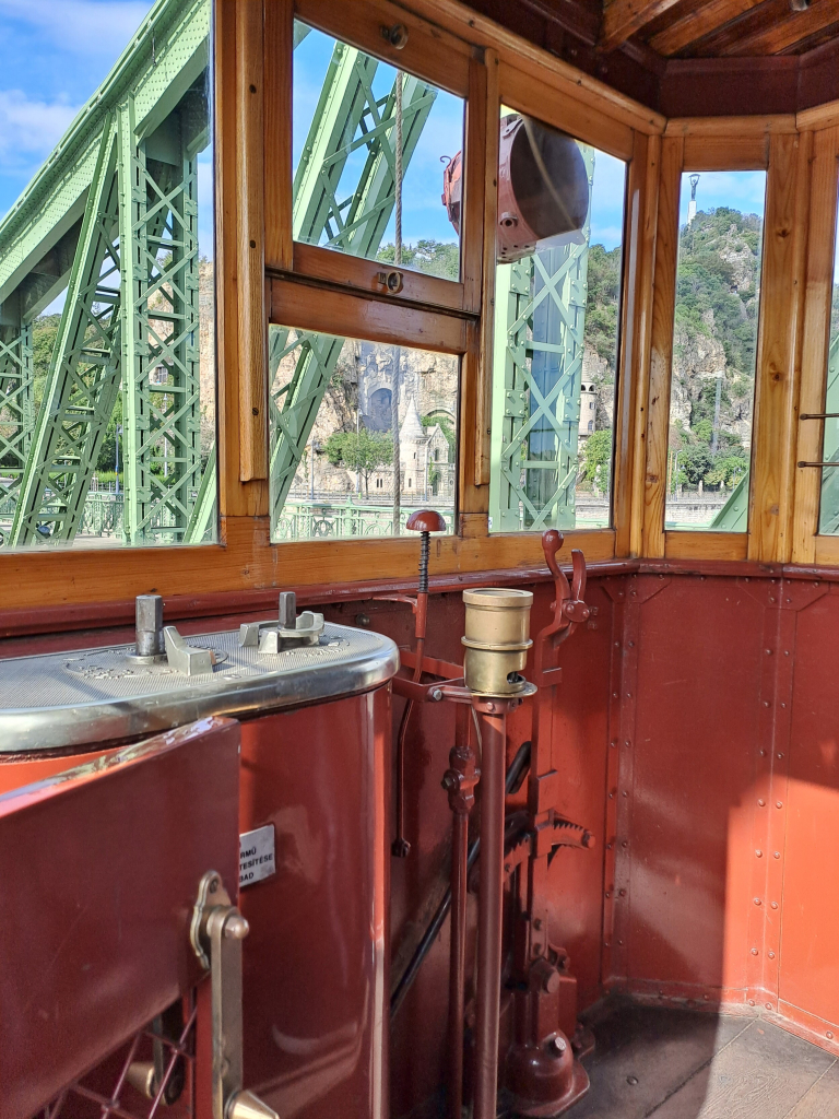 Photo of the inside of the tram, drivers window. Very few parts, one bell button, two hand cranks, wood frames on the windows. Beyond, the bridge and the mountain are visible.