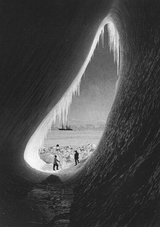 Photograph from inside an ice cave of two men standing in the light of its tear shaped opening, a distant boat in the background.