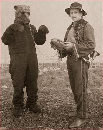 old photo of a man dressed as a bear (kind of unconvincingly) and another man holding him on a leash, wearing a tassled hat, a horn slung over his shoulder and a tambourine in his hands