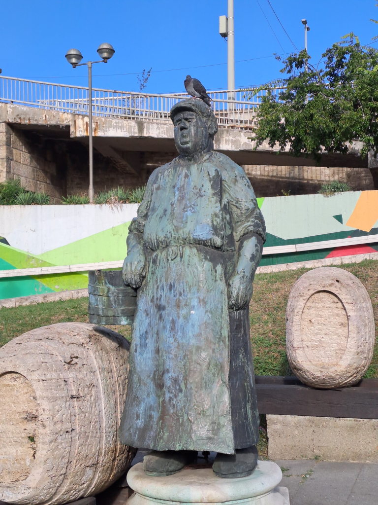 Photo of a weathered (bronze?) statue of an elderly merchant woman at a marketplace. Next to her there are two large barrels carved from stone. She looks tired and worn, staring into the distance. Perched on the statue's head there is a similarly gloomy looking (live) pigeon.