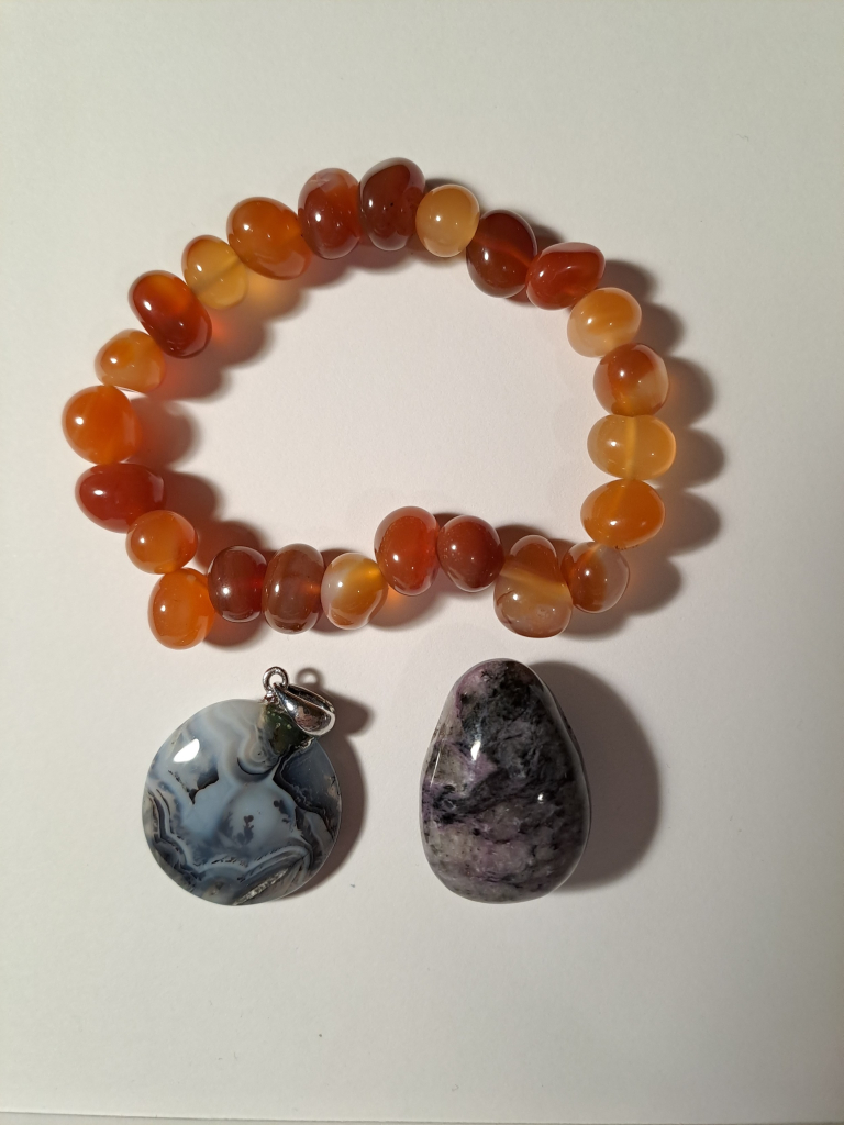 A bracelet made of irregular pebble shaped carnelian beads (about the size of peas), next to a teardrop shaped polished pebble that has white and black patterns with some vivid light purple (charoite) mixed in. The agate pendant is round, marbled black and white, with some branching dendrite features.