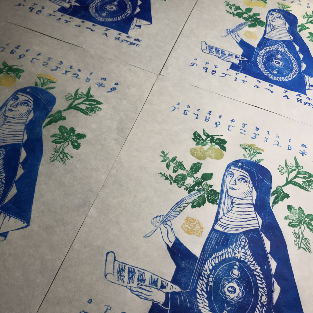 Several of my linocut prints drying on a table. Each one shows Hildegard von Bingen, scroll and quill in hand, dressed in her nun’s habit, printed in blue, with her map of the cosmos, surrounded by green and yellow plants and sulfur. Her invented alphabet runs across the top and bottom. 