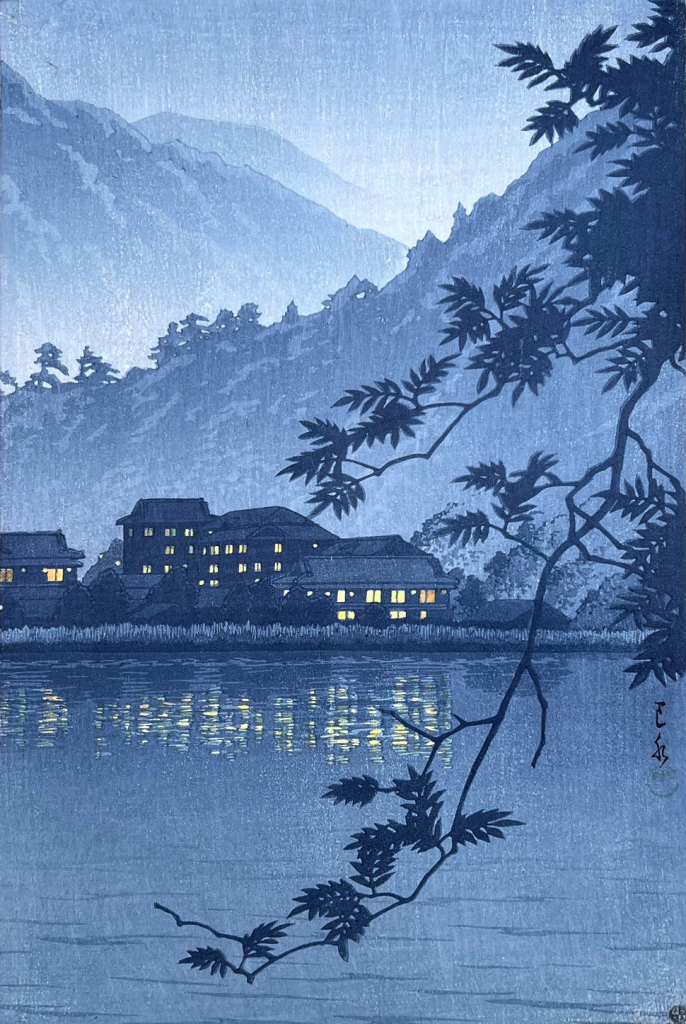 A view looking across Lake Yunoko toward Yumoto Onsen on a rainy evening. In the foreground, silhouettes of tree branches reach out over the the lake. Across the still water, warm lights glow in the windows of Yumoto Onsen; these lights are reflected and scattered on the surface of the lake. Beyond, layers of tree-covered hillside recede into the mist, while above, the sky is hazy and overcast