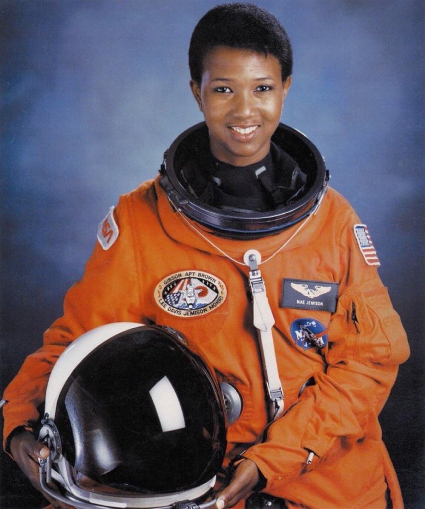 A color photo of Dr. Mae Jemison in her flight suit. The baggy orange suit bears a name tag with a winged insignia, as well as the NASA logo and a flight patch for her mission. She is smiling at the camera. Dr. Jemison is holding the helmet – large and round, with a dark reflective visor – at her side.