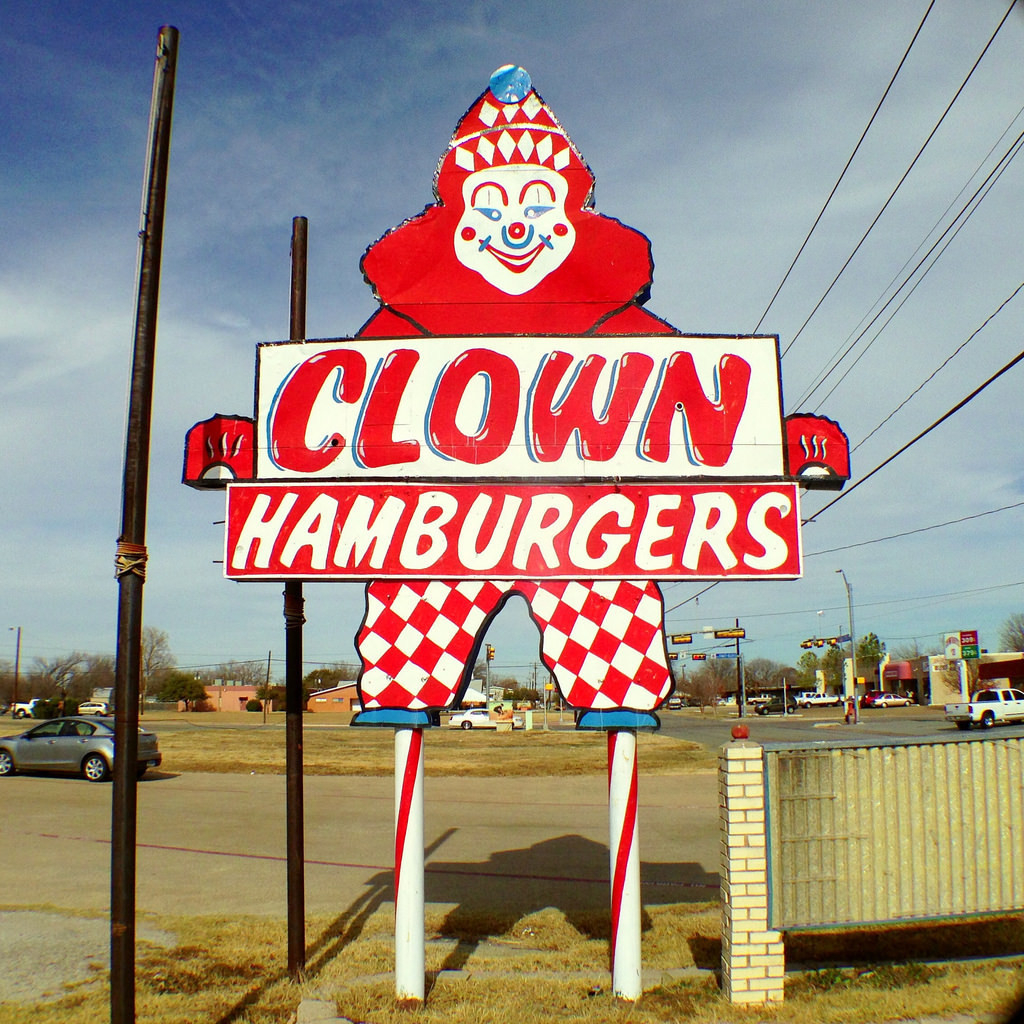 old sign that says: Clown Hamburgers -- the sign is in the shape of a harlequin clown. The clown has a look of sly amusement on its face and is worrying.