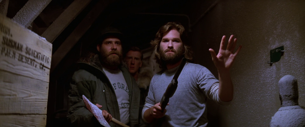 Still from the film "The Thing" (1982). Characters in the antarctic research station advancing towards the camera, armed with an axe and a shotgun. Macready holds up his left hand as if to signal a halt or to calm whatever they are approaching.