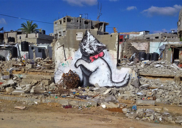 Street art by Banksy on rubble in Northen Gaza. A painting of a cute kitten appears to be playing with a coiled ball of rusted metal on the ground next to the wall, as if it were a ball of wool.