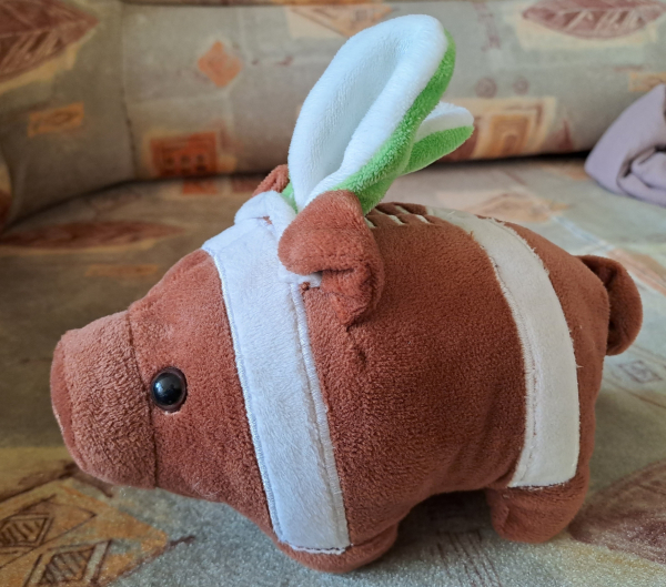 Photo of a plush toy. It's a brown pig, with the shape and stripes of an American football, wearing a headband with white and green bunny ears.