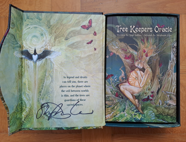 The inside of the open box. Featuring the same art on the art book cover, and a tree and a magpie on the inside lid. The inside lid is signed by Stephanie Law. There is a quote printed on it: As legend and druids can tell you, there are places on the planet where the veil between worlds is thin, and the trees are guardians of these sacred spaces.