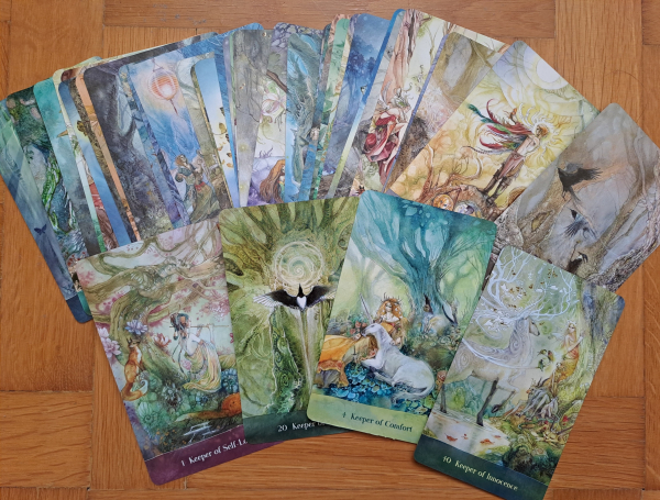 Colorful cards from the deck, fanned out. Visible are Keeper of Innocence (a fairy like woman peeking from behind a tree at a radiant white stag with branching antlers), Keeper of Comfort (a lady with a unicorn resting on her lap under a tree), Keeper of Self-love (a woman playing a flute surrounded by forest animals).