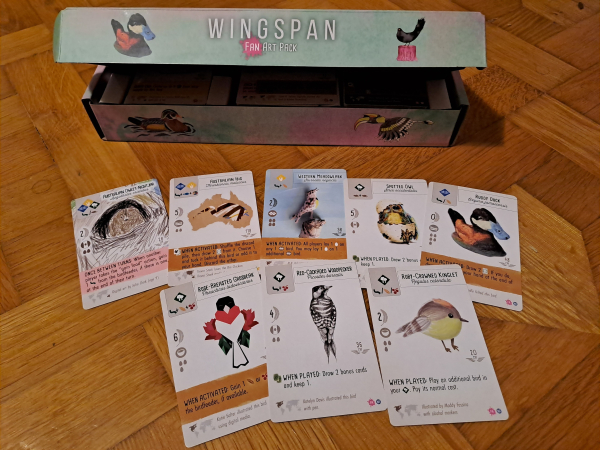 Open Wingspan box with some cards spread out in front of it. Images include a kid's color pencil drawing of an owl, a needle felted duck, a clay meadowlark, and an adorably spherical kinglet 