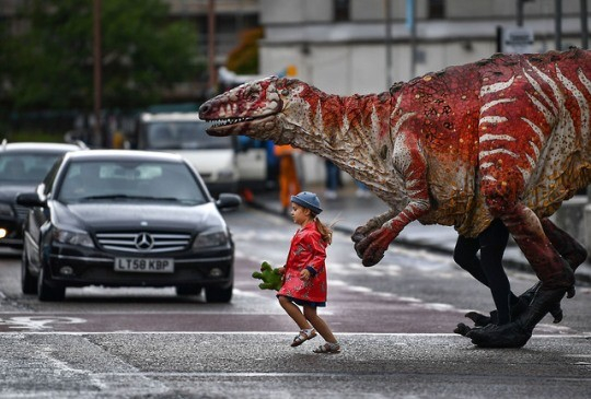 a little girl in a red raincoat and blue hat, holding a teddy, crosses the street with her majungasaurus -- a carnivorous dinosaur that is red and white