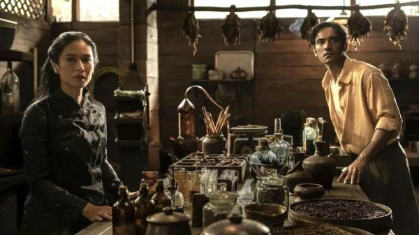 Dian Sastrowardoyo and Ario Bayu stand at a table covered with bottles and flavorings for spice cigarettes.