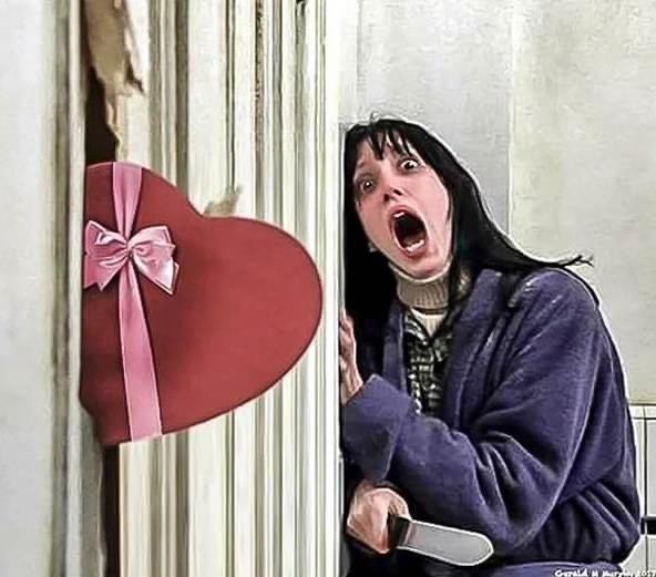 iconic still of shelly duval screaming in The Shining, but instead of an ax breaking through the door, it's a heart-shaped box of chocolates