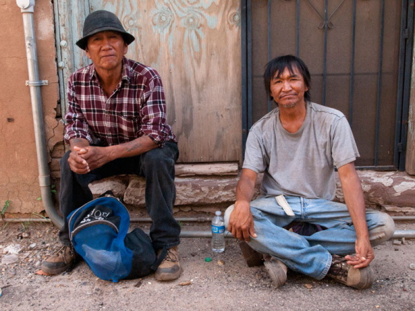 Two men, one Hopi, wearing a plaid shirt, felt hat, jeans, and sneakers with a day pack between his feet, and one Navaho, wearing a t-shirt and jeans with a water bottle to his left. They are sitting in front of a door that is half boarded up and half security screen in an alley in Winslow, Arizona. Neither Jackson Browne nor a girl in a flatbed Ford were anywhere to be seen.