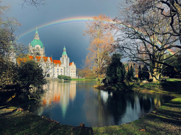 The old bright Hanover town hall which looks a bit like a castle is in the background on the left under a dark grey sky with a fantastic rainbow above. A beautiful park landscape with a round stone path and a lake can be seen in the front. The shape matches the rainbow's bow, so it looks like a connected oval. There are trees and bushes on both sides of the little lake and light reflections everywhere. The town hall is also reflected in the lake.