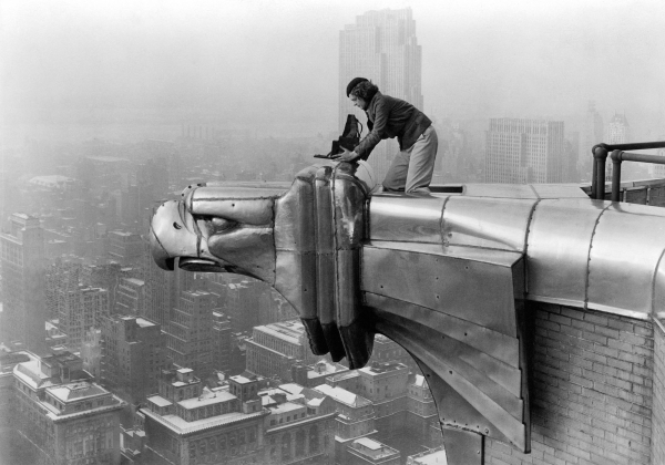 Black and white photo of Margaret in the 1930s, positioning her camera while kneeling on an eagle statue on the 61st floor of the Chrysler Building, high above the skyline.