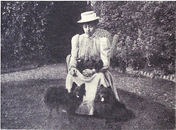 Black and white photo of Sophia, sitting in a garden in a white, closed-neck Victorian dress, wearing a hat with her hair pinned up. She has a serious expression on her face. She is surrounded by three black pet dogs (I am bad at dogs but they look like Pomeranians)