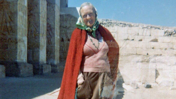 Photo of Omm Sety in front of the Abydos temple, in her elderly years. She is wearing a pink vest, brown pants, a red blanket over her shoulders, and a green head scarf. She is smiling cheerfully into the camery.