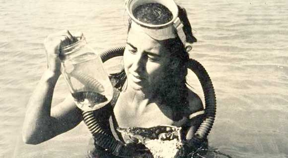 Black and white photo of a young Eugenie in diving gear, chest-deep in water and holding a glass jar with some small striped fish in it.