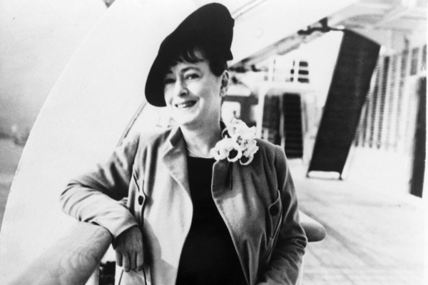 Black and white photo of Dorothy Parker. She is smiling and leaning on a balustrade, wearing a light coat, and a hat at a jaunty angle.