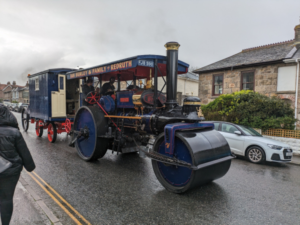 A blue vintage steamroller pulling a wagon down a street. 