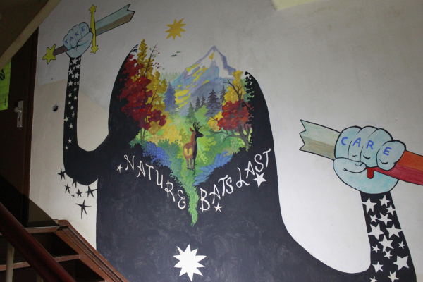 A mural painting in Kanthaus stairs, depicting a piece of autumnal wilderness, with a stag in the woods at the based of a mountain. The whole landscape forms an anthropomorphized character who broke a bloody sword with its hands. The character's hands display the words "Take care", and its trunk "Nature bats last".