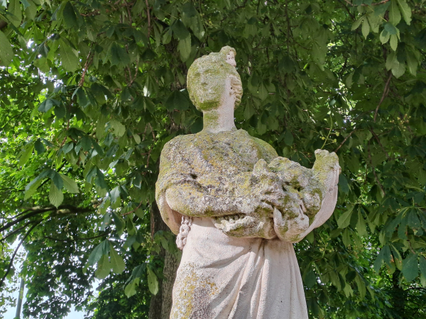 Photo of a statue depicting a woman who carries flowers (fruit?) in the folds of her dress. The statue is weathered, face almost blank, covered in moss and lichen. There is a big chestnut tree in the background. 