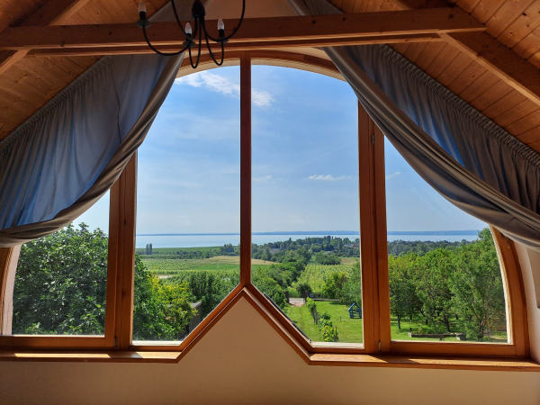 A large semicircular window framed by wooden frames and the beams of a wooden roof. Light blue curtians are drawn to the side. The view from the window is a sunlit landscape of vineyards, orchards, trees, and bright blue Lake Balaton in the distance under a summer sky.