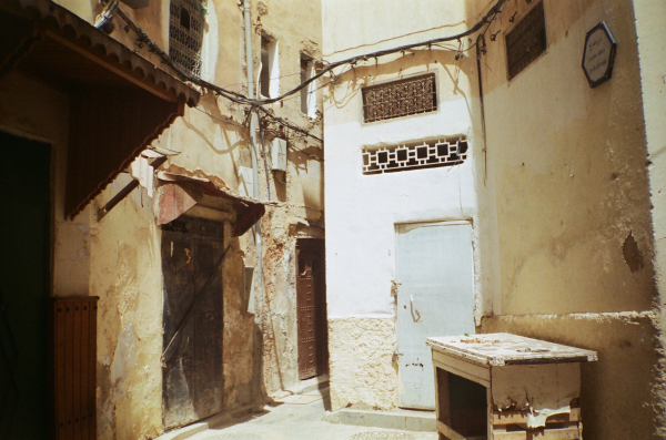 Narrow, sunlit alley with aged, textured walls and several small doors. The wiring runs across the upper walls. A worn table stands against a wall.