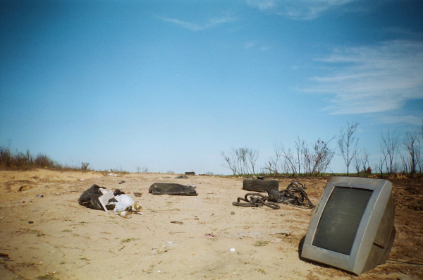 A deserted sandy area with scattered trash, including plastic bags, tires, and an old CRT monitor, under a clear blue sky. Sparse, dry vegetation is visible in the background.