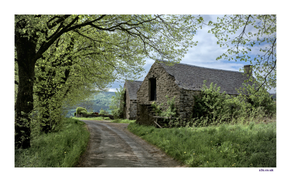 A small country road curves past an old stone barn. There are trees, bushes and grass beside the road. The sky behind the barn is blue with some white cloud. The photo feels warm and sunny.