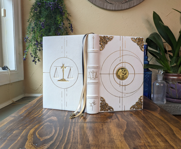 A white leather journal standing upright on a deck. The journal is open with the spine towards the viewer to show both covers at once. It has a gold design on both covers.