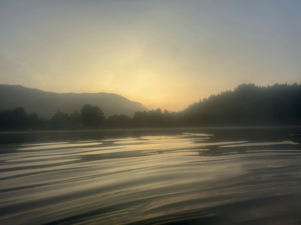 Looking over the water towards a gap in the hills and lochside trees where the golden light of the unrisen sun is glowing in a misty sky. The glassy water in the foreground is rippled by my swimming.