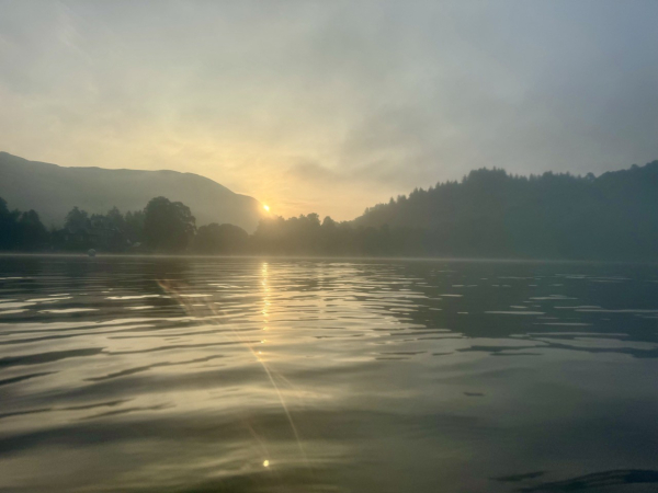 Misty view across a loch the golden sun appearing from behind monochrome hills on the left and shining a trail across the water. 