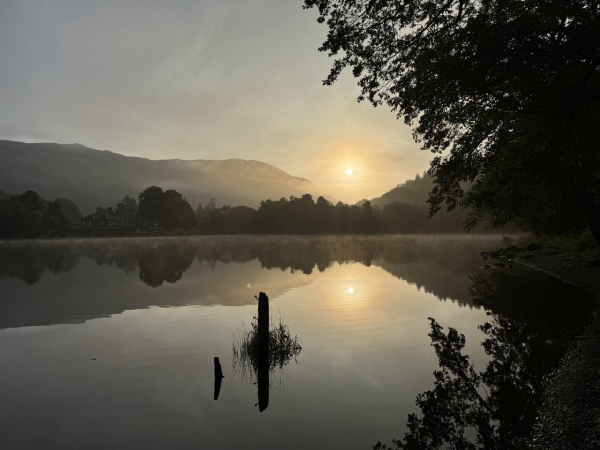 Perfect reflections on a loch of a pale sky and a hazy golden sun gleaming though a gap in the grey hills and black trees on the shore.