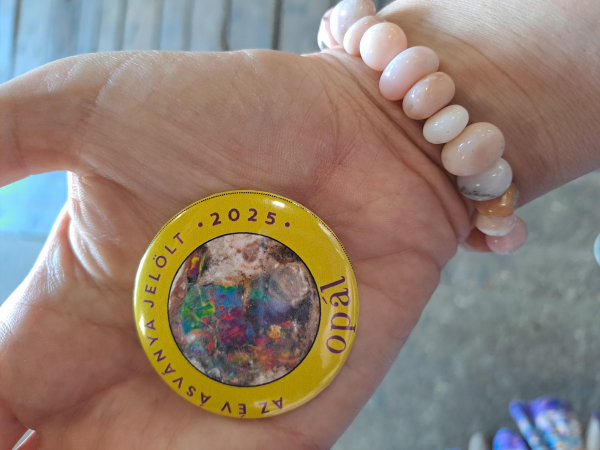 Up close photo of my hand, holding a button on my palm. Button has a photo of a colorful piece of opal, and a yellor border that reads "Mineral of the Year Candidate 2025 - Opal". I am also wearing a bracelet of pale pink opal pebbles.