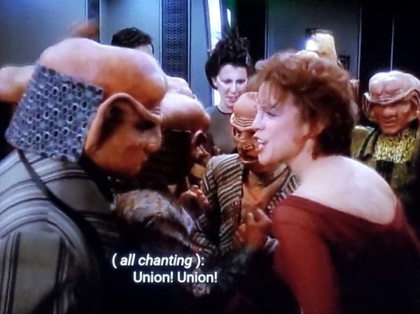 DS9 scene. A humanoid Bajoran (Leeta) is standing in the middle of a group of Ferengi. She looks super pumped up and is cheering them on. Closed caption reads, "(all chanting): Union! Union!"