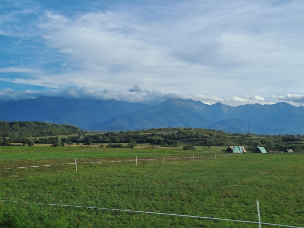 Scenic photo of green fields, low wooded hills, and tall mountains in the backdrop with their peaks covered in clouds. A fence and hay bales are visible in the field.