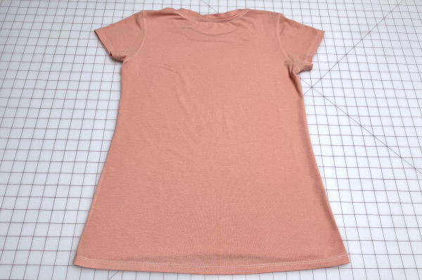 Rear exterior view of a freshly sewn peach jersey t-shirt laying flat on a large cutting mat.