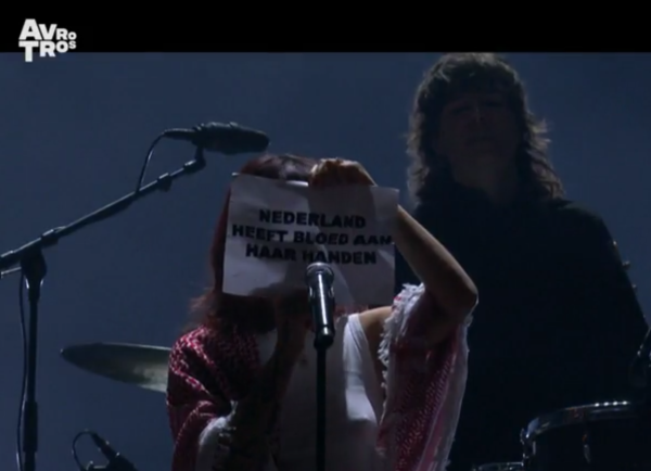 Uitsnede van een screenshot van de live-uitzending van het openingsconcert van 750 jaar Amsterdam met Sophie Straat die een stuk papier met de tekst "NEDERLAND HEEFT BLOED AAN HAAR HANDEN".