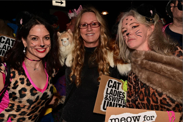 3 women dressed up. 2 on the outside in leopard print with cat makeup. One in the middle is wearing a black leotard with life-size cats around her neck.