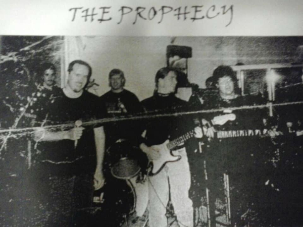 An old heavily-creased faded black and white photograph of a band of five members. I am front and centre holding a guitar and my fly is down. The band name is written above the picture and reads: THE PROPHECY 