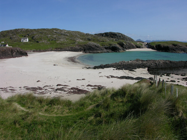 The beach and coastal scenery at Clachtoll. The image shows a view over a broad white beach at low tide from tussocky grass in the foreground to a rocky outcrop on the right and a low rocky promontory on the far side of the beach, with some white cottages at its left-hand end. The turquoise sea is lapping in from the right and in the background are some mountains low on the horizon on the right of the frame. The sky is blue.