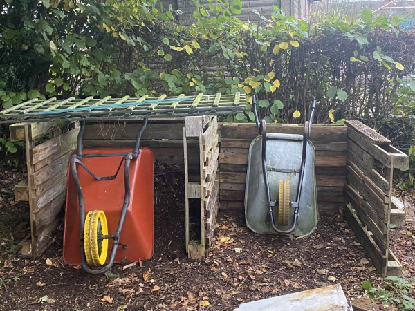 A two bay composting area made out of reclaimed wooden pallets. The left hand bag already has compost material in with a red wheelbarrow leaning against it. The right hand bay is empty except for a small silver barrow leaving against the back. 